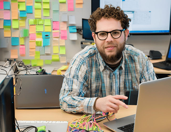 An IT guy wearing eyeglasses. The IT person's glasses reflect the bright laptop screen, their desk cluttered with gadgets, sticky notes, and a neatly coiled ethernet cable.